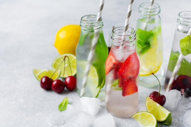 Un montón de botellas con refrescante limonada de verano con limón, fresa, cereza, pepino y hielo sobre un fondo de hormigón gris