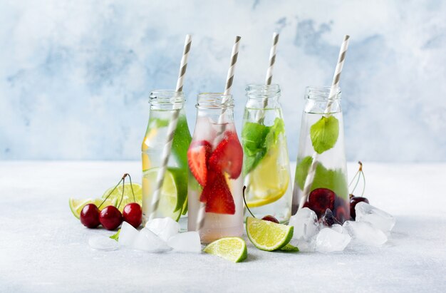 Un montón de botellas con refrescante limonada de verano con limón, fresa, cereza, pepino y hielo sobre un fondo de hormigón gris.