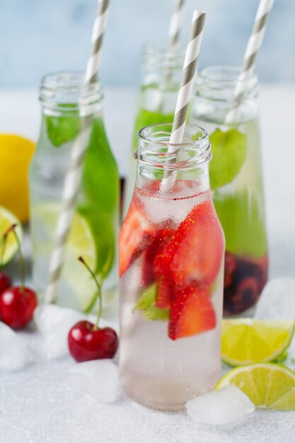 Un montón de botellas con refrescante limonada de verano con limón, fresa, cereza, pepino y hielo sobre un fondo de hormigón gris.