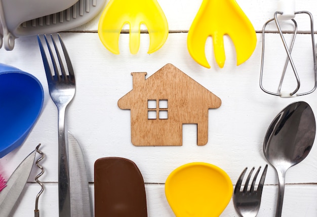 Un montón de diferentes utensilios de cocina en la mesa de madera y una pequeña casa de madera entre ellos.