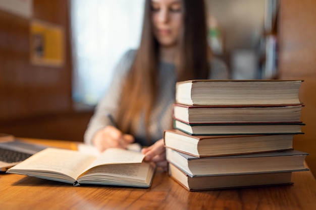 Montón de libros sobre la mesa en la biblioteca Joven estudiante morena concentrada escribiendo en el cuaderno y trabajando con un libro en el fondo