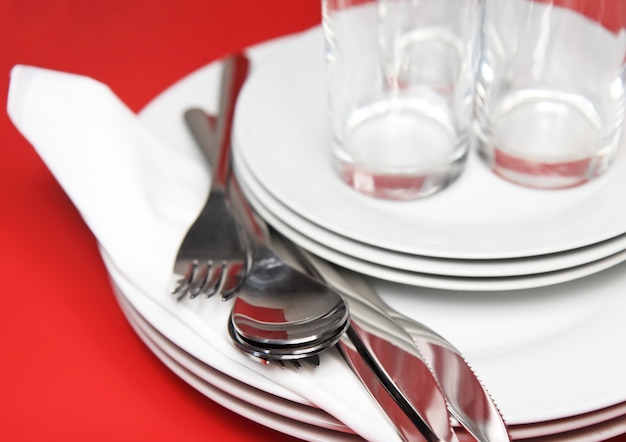 Montón de platos blancos, vasos con tenedores y cucharas sobre una servilleta de seda. Fondo rojo