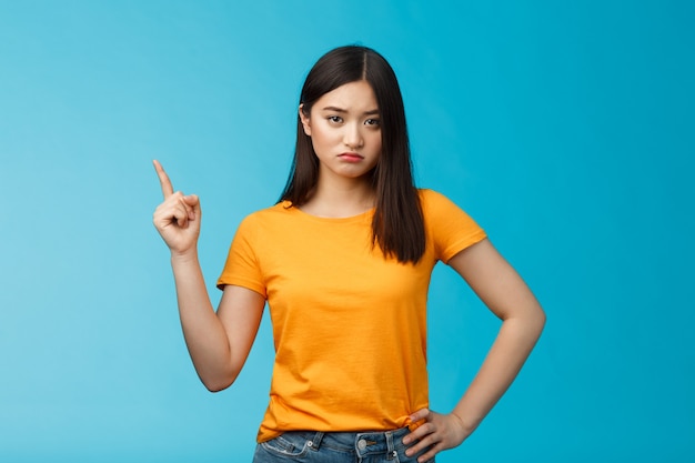 Moody verärgerte asiatische Freundin, schmollender Blick beleidigt und unzufrieden, die obere linke Ecke zeigt, enttäuscht schmollend, stehender beunruhigter blauer Hintergrund tragen gelbes T-Shirt. Platz kopieren