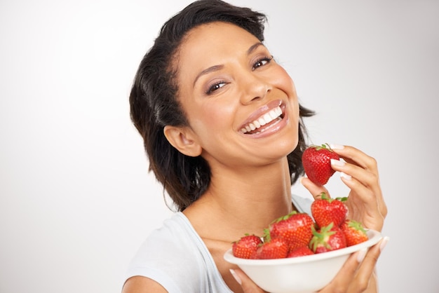 Foto morangos saudáveis e uma jovem em um estúdio para nutrição de bem-estar e dieta orgânica vitaminas de sorriso e retrato de uma pessoa feminina comendo uma fruta para um lanche vegano saudável por fundo cinza
