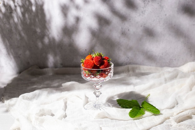Morangos vermelhos frescos em um copo sob a sombra de flores sobre uma mesa em um fundo de concreto.