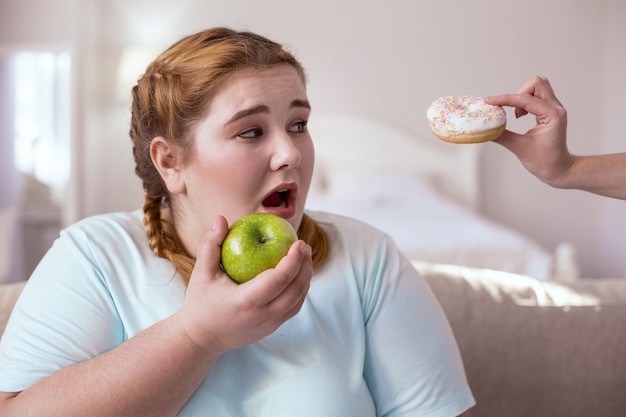 Una mordida. Mujer agradable regordeta que decide probar un bocado de rosquilla mientras sostiene la manzana verde en la mano