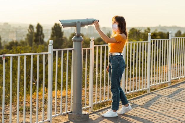 Morena acercándose a los binoculares en un parque público mientras usa una máscara quirúrgica.