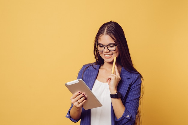 Morena adorable en gafas con el pelo largo vestido con traje azul oscuro linda sonrisa, sosteniendo su tableta, se ve feliz, hace videollamada, sobre fondo amarillo con espacio de copia. Las personas sinceras emociones.