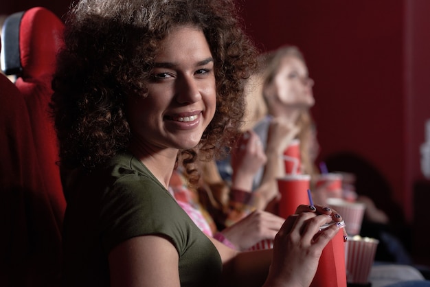 Morena alegre com um lindo sorriso, comendo pipoca durante a exibição de um filme interessante no cinema.