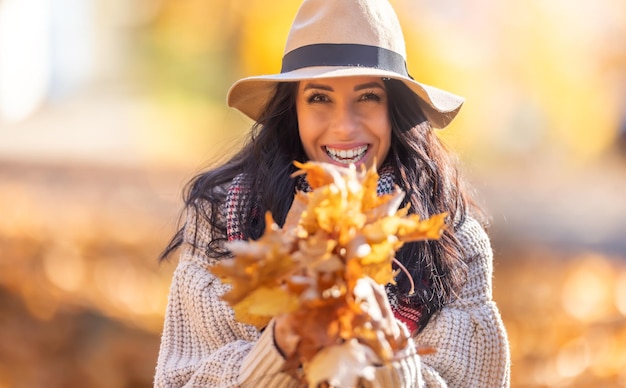 Una morena alegre con ropa elegante de otoño sostiene un brazo lleno de coloridas hojas caídas