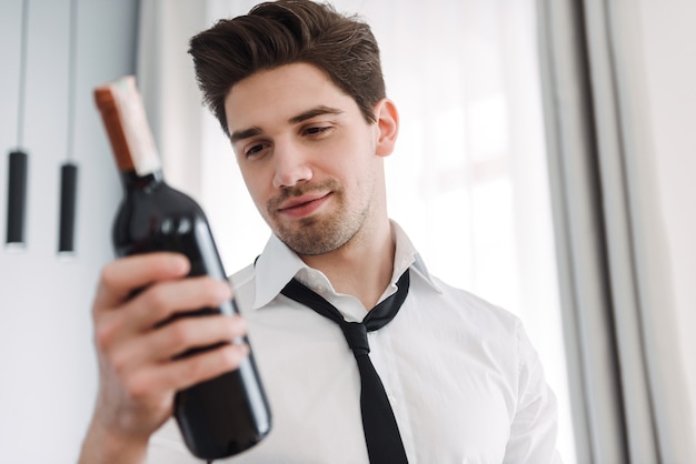 Morena barbudo empresario vistiendo ropa formal sosteniendo una botella de vino en el apartamento del hotel