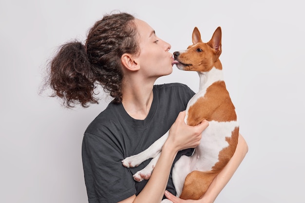 Morena beija a cadela basenji apaixonada por seu animal de estimação favorito expressa seu cuidado vestido com uma camiseta preta isolada sobre o branco