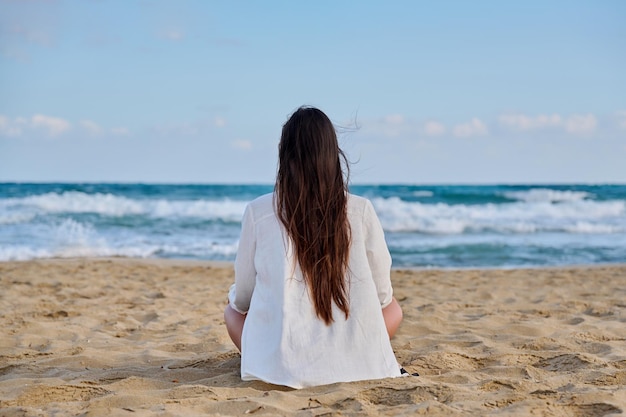 Morena de blanco con el pelo largo sentada en la playa de arena con la espalda mirando al mar