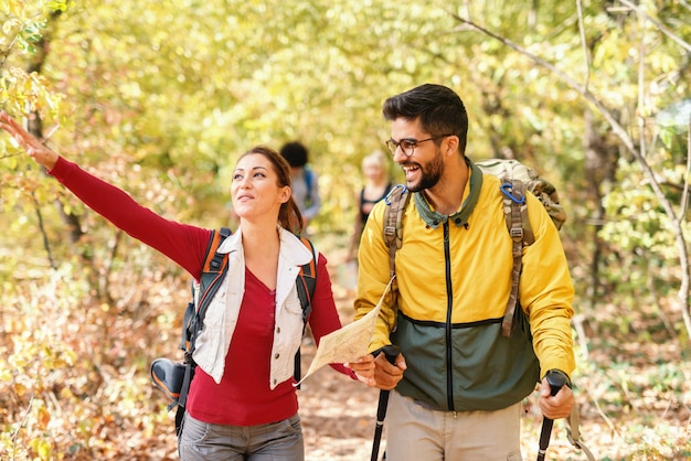 Morena bonita de sorriso que guarda o mapa, mostrando o caminho certo e conduzindo o resto dos caminhantes. ao lado dela, andando barbudo homem sorridente com óculos. floresta no exterior do outono.