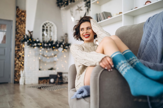 Morena bonita e feliz sentada na cadeira com um agasalho quente e luzes de natal atrás