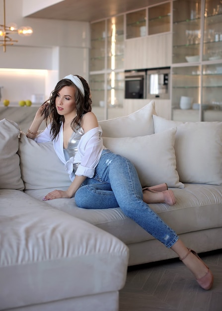 Foto morena bonita no sofá em uma camisa branca, calça jeans e sapatos bege. retrato de uma menina em um interior luminoso.