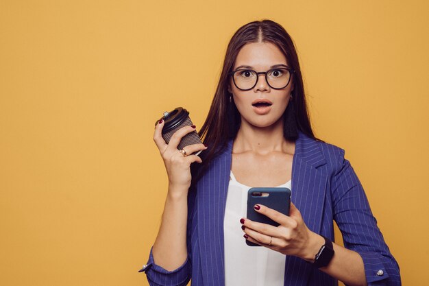 Morena caucasiana chocada de óculos e terno segurando o telefone em uma mão e xícara de café em outro tendo o rosto surpreso