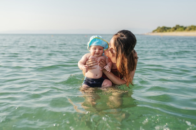 Morena caucasiana comprometida ensinando seu amado filho de 6 meses a nadar no mar. Bebê com chapéu na cabeça, curtindo e sorrindo
