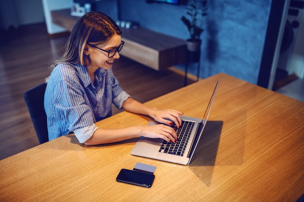 Foto morena caucásica hermosa joven con las lentes que se sientan en la mesa de comedor y que usa la computadora portátil.