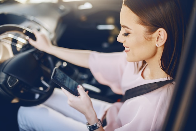 Morena caucásica joven atractiva con sonrisa dentuda y vestido elegante coche de conducción y mirando el teléfono inteligente.