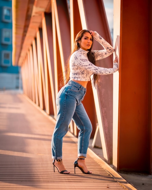 Una morena chica latina en jeans en la ciudad al atardecer en un suéter naranja