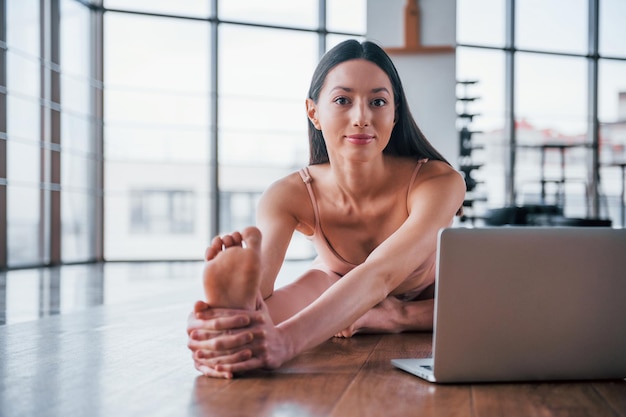 Foto morena com corpo magro fazendo exercícios no chão e usando o laptop.