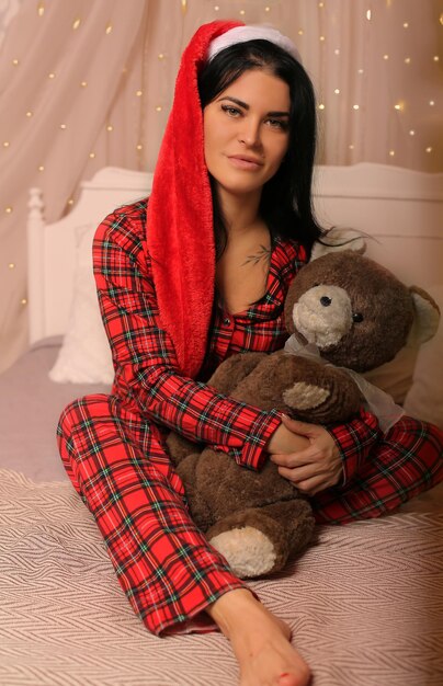 morena com pijama de Natal chapéu de Papai Noel sentada na cama abraçando um ursinho de pelúcia