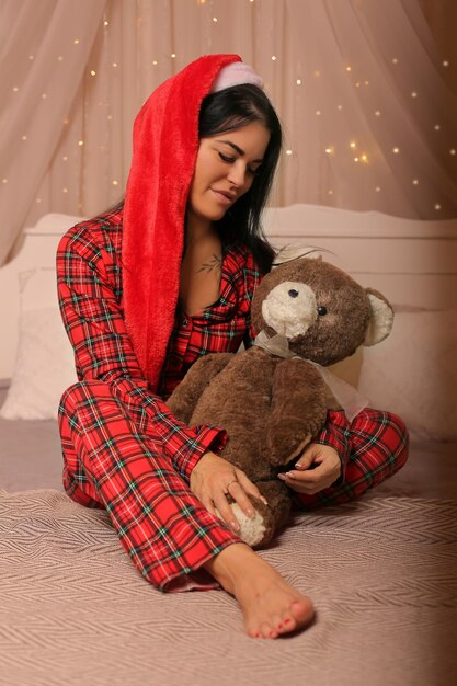 morena com pijama de Natal chapéu de Papai Noel sentada na cama abraçando um ursinho de pelúcia