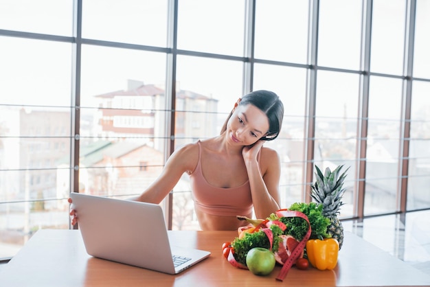 Morena com tipo de corpo magro senta-se à mesa com laptop e comida saudável.