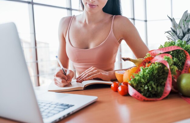Morena com tipo de corpo magro senta-se à mesa com laptop e comida saudável.