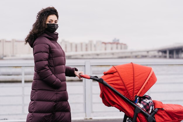 Morena com uma máscara médica, vestida com uma jaqueta quente na rua contra a cidade, rola um carrinho de bebê na frente dela na cidade.