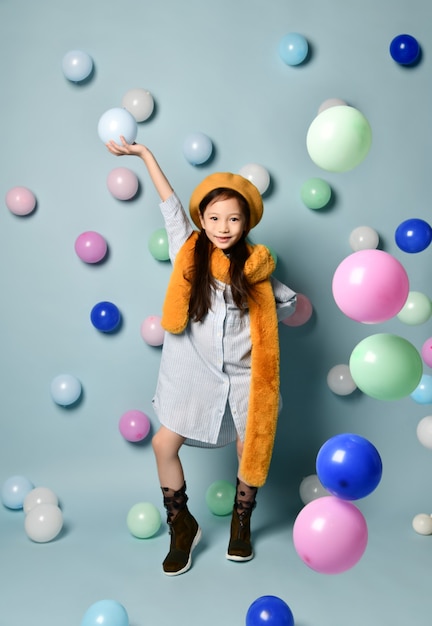 Morena criança asiática em vestido de camisa grande, boina marrom e cachecol, botas. Ela sorrindo, posando com balões multicoloridos sobre fundo azul. Infância, moda, estilo moderno. Comprimento total