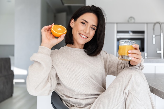 Morena de 30 anos bebendo suco de laranja enquanto descansa em um quarto moderno e bem iluminado