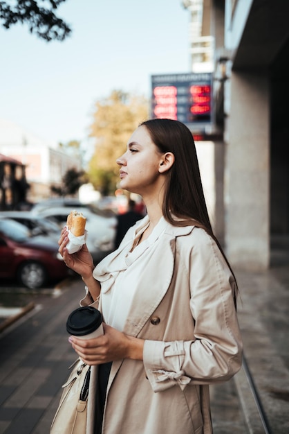 Morena de cabelos compridos com um casaco leve está comendo bolo e café em uma rua europeia