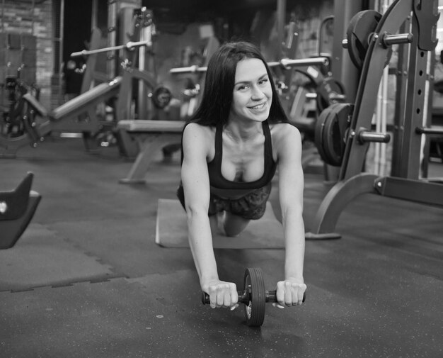 Morena de deportes Smilling en ropa deportiva haciendo ejercicio con rodillo de abs en el gimnasio y mirando a cámara.