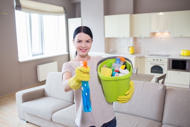 Morena em pé e posando na câmera. ela segura um frasco de spray azul e um balde com equipamento de limpeza. ela sorri e mira na câmera.