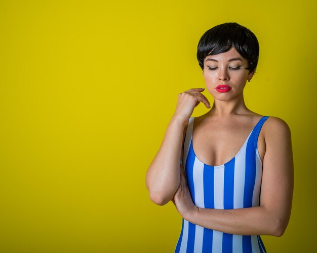 Morena em um maiô listrado posando no estúdio em um fundo amarelo brilhante Mulher com uma peruca curta com lábios sensuais