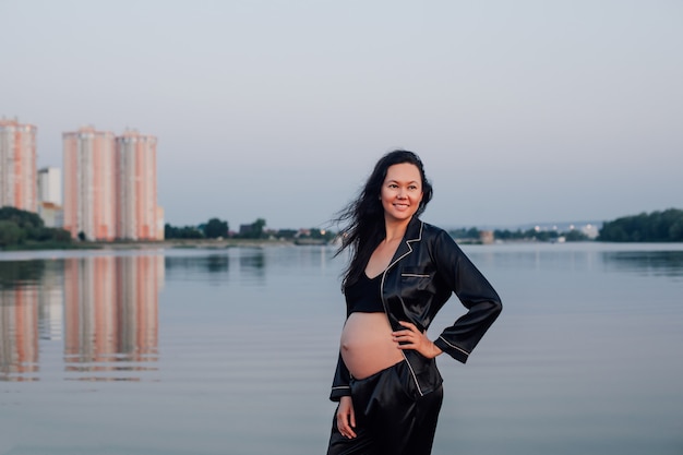 Morena embarazada sonriente cerca del agua joven hermosa mujer sexy vestida con traje oscuro de seda plantea agai ...
