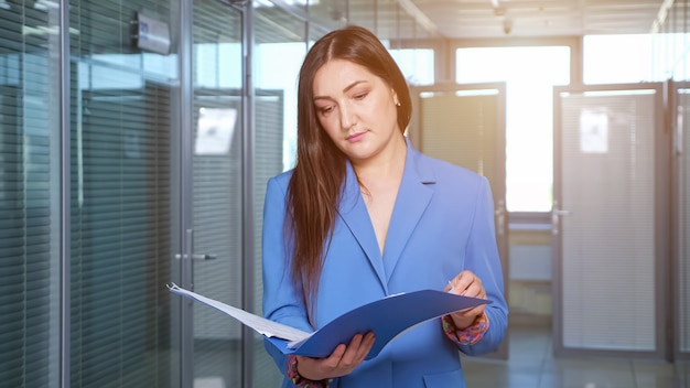 Morena empresaria concentrada mira a través de hojas de informes de la empresa en una carpeta azul de pie en el pasillo de la oficina contra las ventanas, la luz del sol