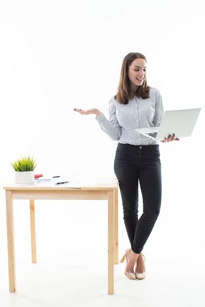 La morena está de pie junto a la mesa y trabaja en una computadora sobre un fondo blanco.