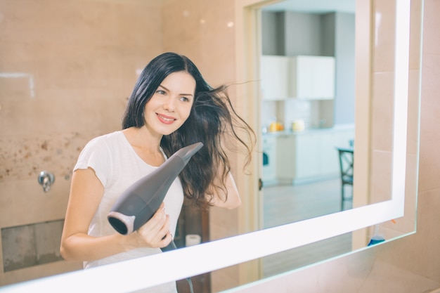 Morena está sentada en el espejo del baño y la mira. Ella se está secando el pelo. La mujer sonríe Ella se ve positiva.