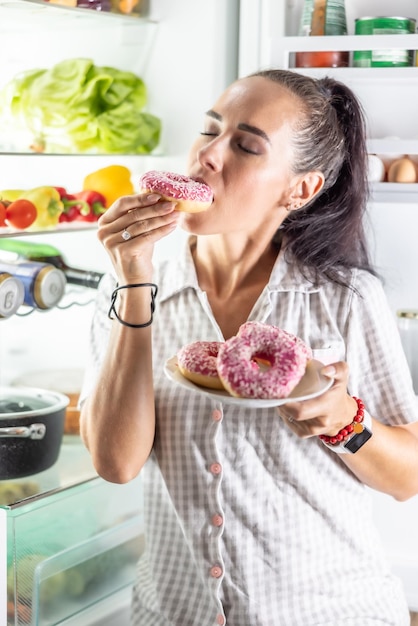 Foto morena faminta de pijama desfruta de donuts doces tarde da noite perto da geladeira aberta