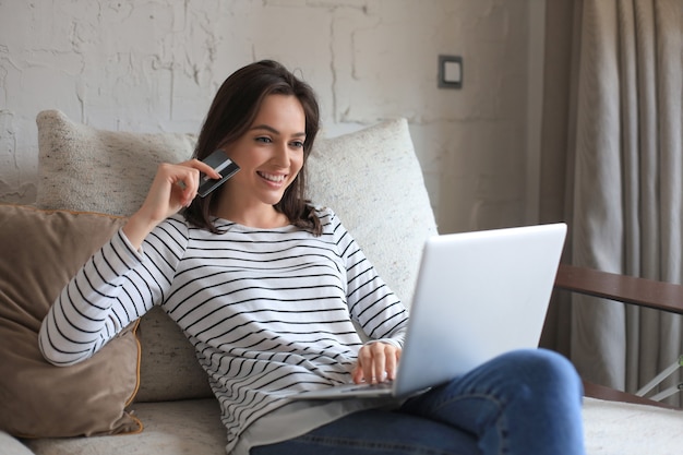 Foto morena feliz natural usando cartão de crédito e laptop na sala de estar.