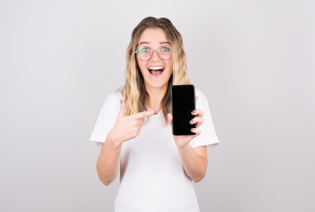 Morena feliz sorprendida en chaqueta de mezclilla que muestra la pantalla del teléfono inteligente en blanco y apuntando hacia ella, alegremente con la boca abierta en la pared blanca