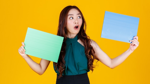 Morena feminina asiática de cabelo bonito e muito encaracolado segurando poses de placa em branco verdes e azuis para a câmera com uma alegria para fins de uso de publicidade, estúdio tiro isolado em fundo amarelo.