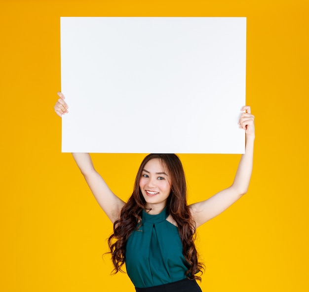 Morena feminina asiática de cabelo bonito e muito encaracolado segurando uma placa branca em branco sobre a cabeça com uma alegria para fins de publicidade e banner uso, estúdio tiro isolado em fundo amarelo brilhante.