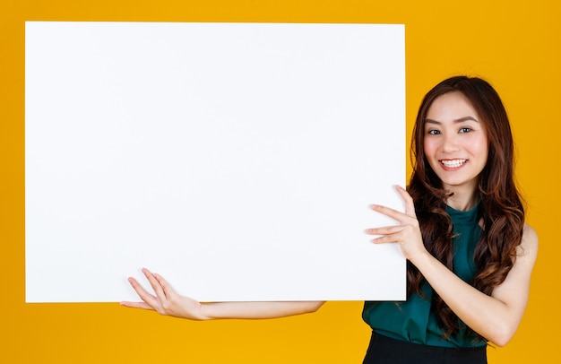 Morena feminina asiática de cabelo bonito e muito encaracolado segurando uma placa em branco branca posa para a câmera com uma alegria para fins de publicidade e banner, tiro do estúdio isolado no fundo amarelo brilhante.