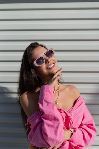 Morena hermosa joven en un collar de joyería de cuello de camisa rosa gafas de sol de moda en el fondo de una valla de puerta de garaje de luz