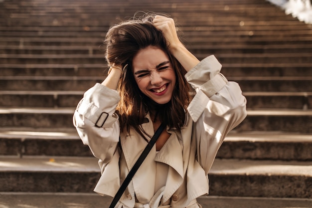 Morena jovem alegre com capa impermeável bege posando, fechando os olhos, balançando o cabelo com as mãos e se divertindo no muro das escadas da cidade velha ao ar livre