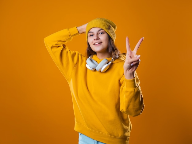 Morena jovem e elegante, alegre, com um moletom amarelo e um chapéu, mostra um gesto de vitória com dois dedos ...